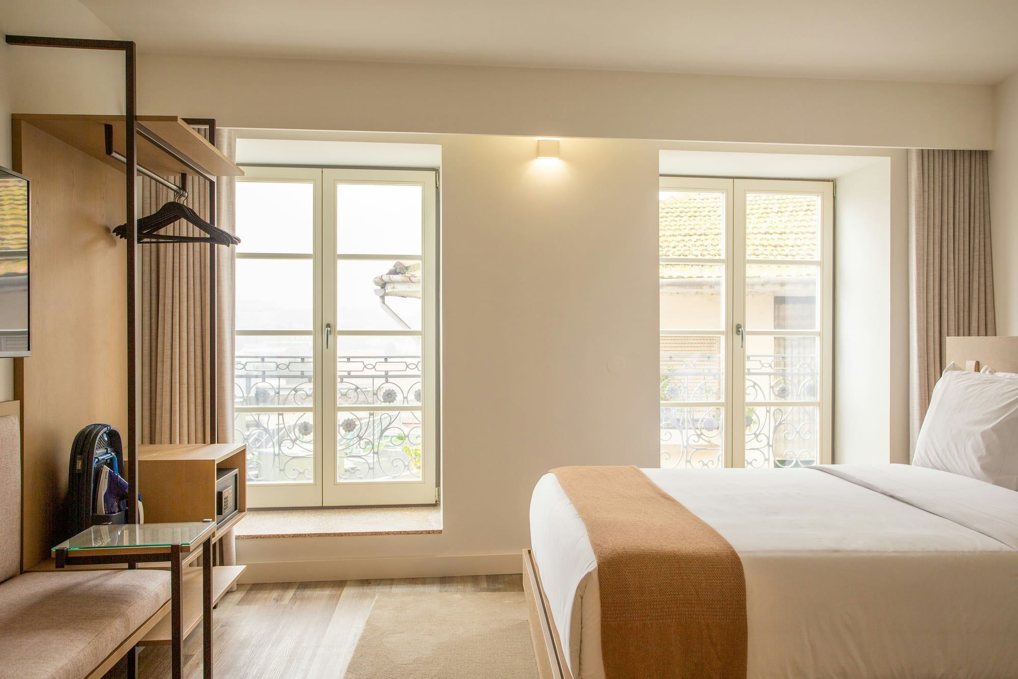 A freshly made hotel room with a double bed and tall windows with iron railings
